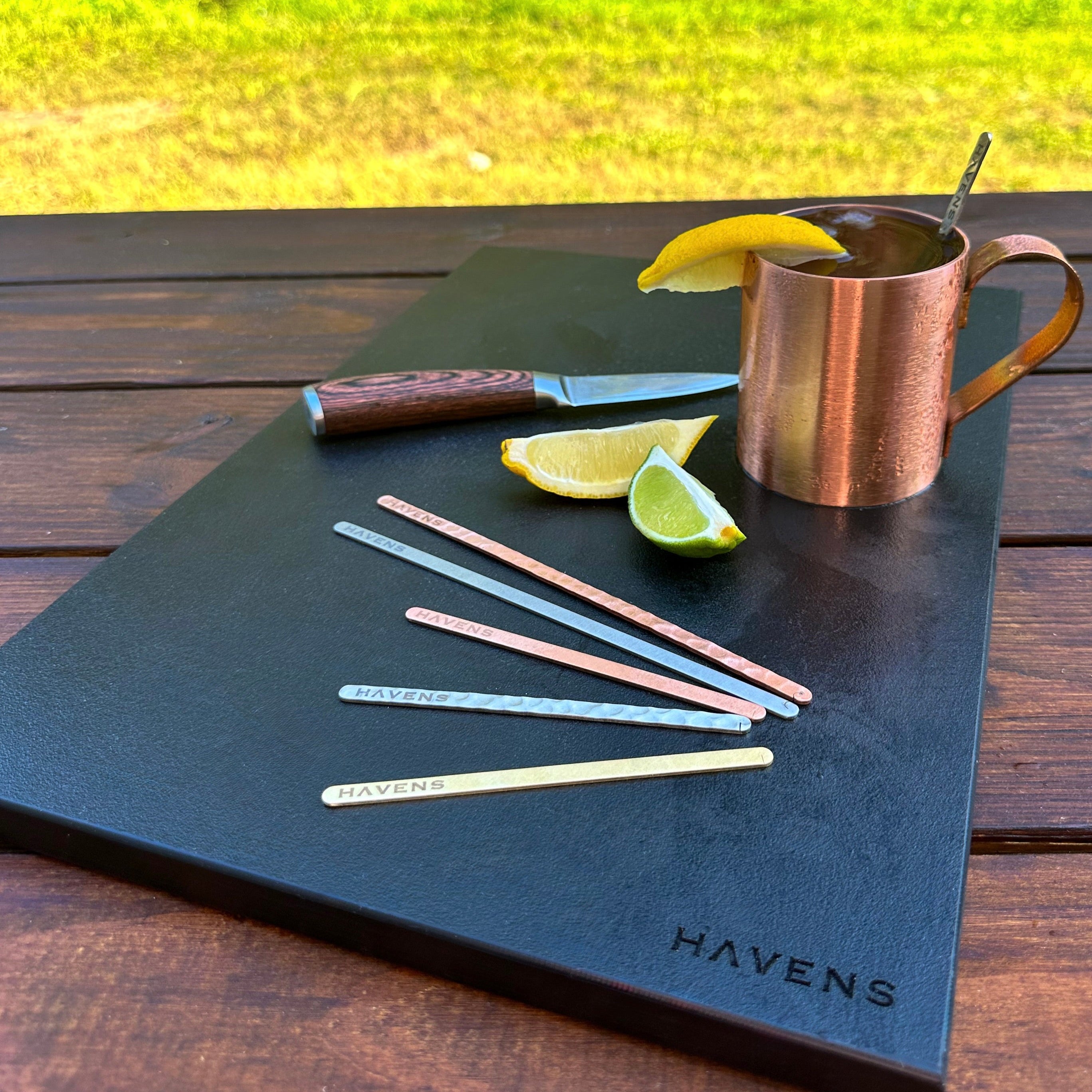 Copper mug with lemon slices on a black mat with 'HAVENS' branding, outdoors.