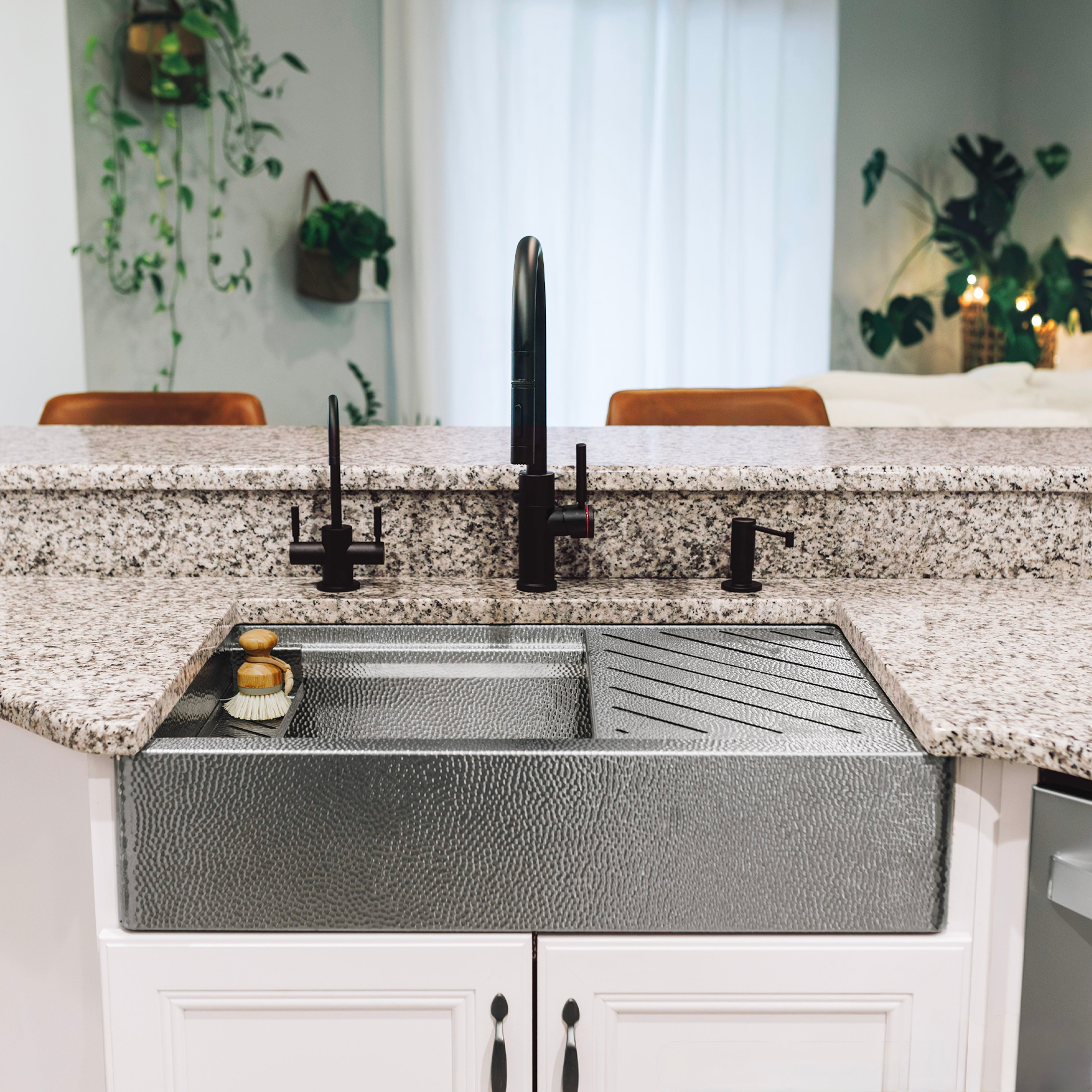 A micro hammered stainless steel farmhouse sink with a matching sponge caddy and drying rack, set into a speckled granite countertop with a black faucet and handles in a bright kitchen. Made in USA