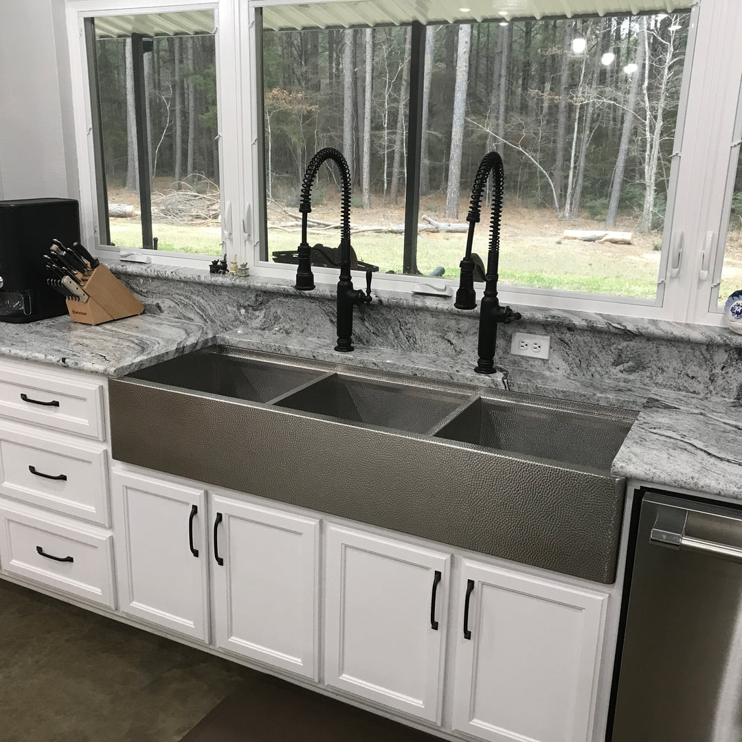 Modern kitchen with a large triple bowl micro hammered stainless steel farmhouse sink, black faucet, and white cabinets.