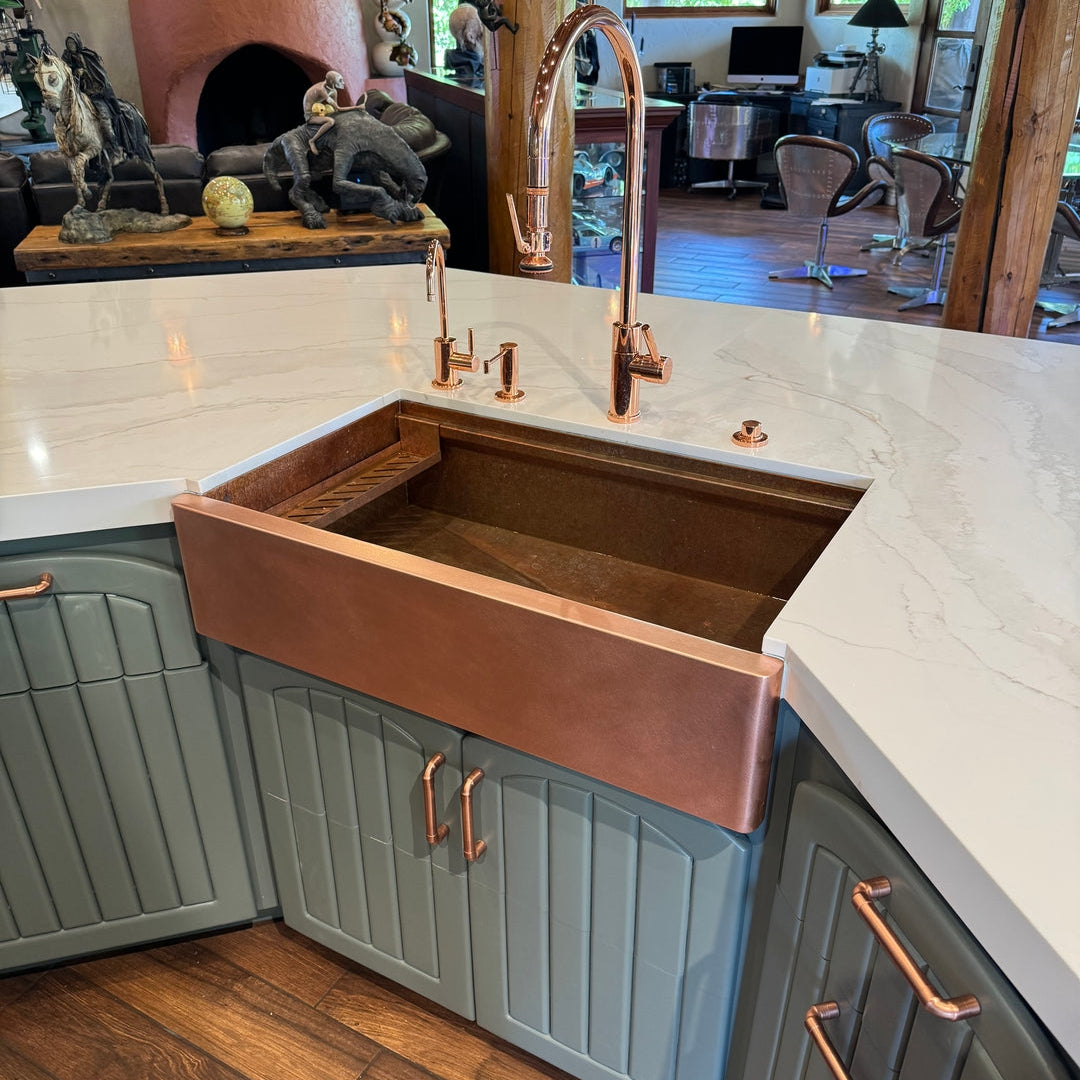 Copper workstation farmhouse sink in a kitchen setting with blue cabinets and wooden flooring.