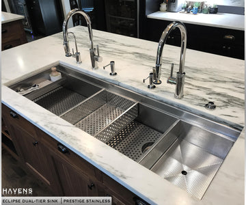 Eclipse dual tier sink with white marble countertops and two faucets featuring custom basin grate, drop in strainer, drop in bowl, and sponge caddy.